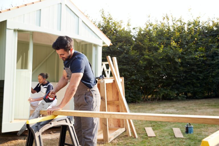 Male Carpenter With Female Apprentice Sawing Wood To Build Outdoor Summerhouse In Garden